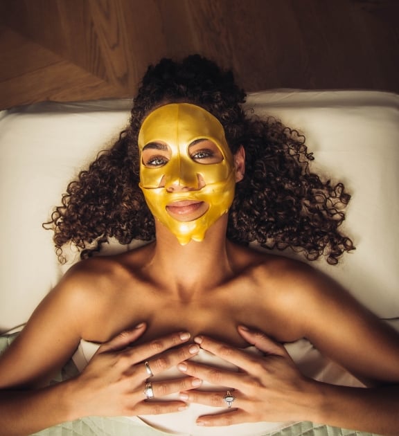 A woman lies down with a gold face mask on her face.