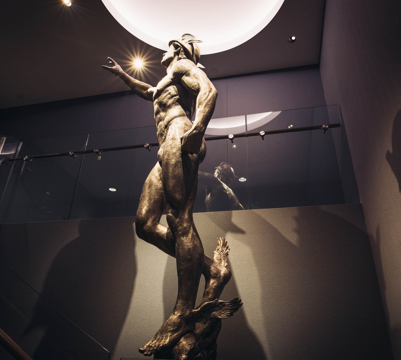 A bronze statue of a man with outstretched arms stands in a room with a glass railing and a circular light on the ceiling.