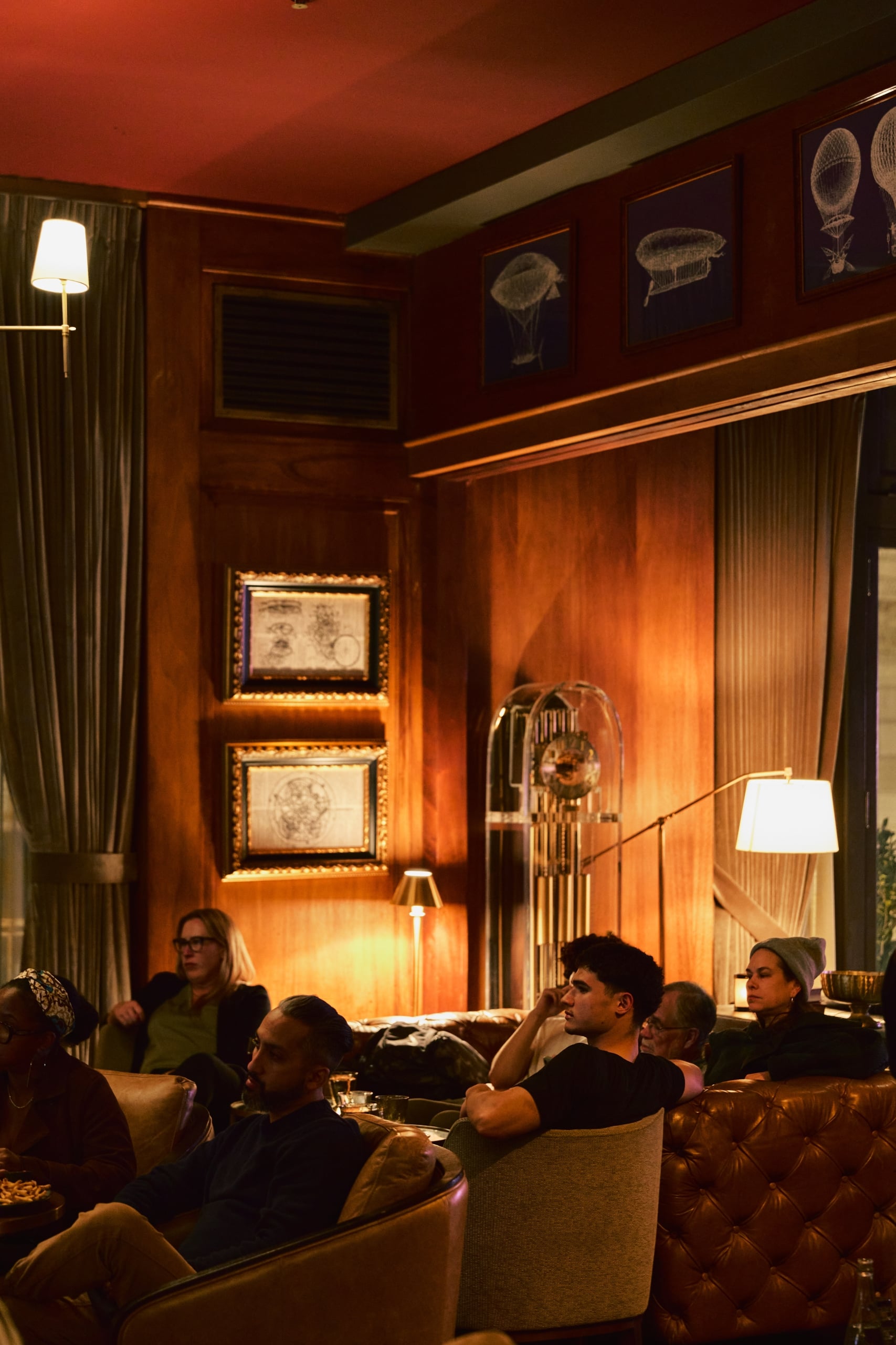 Guests relax in leather armchairs inside a warmly lit lounge with wood-paneled walls, framed art, and vintage floor lamps.