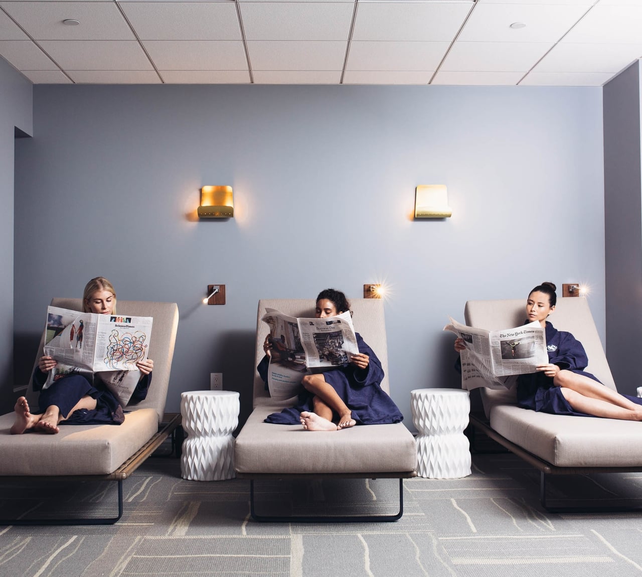 Three women in robes sitting in a row reading newspapers in a spa lounge.