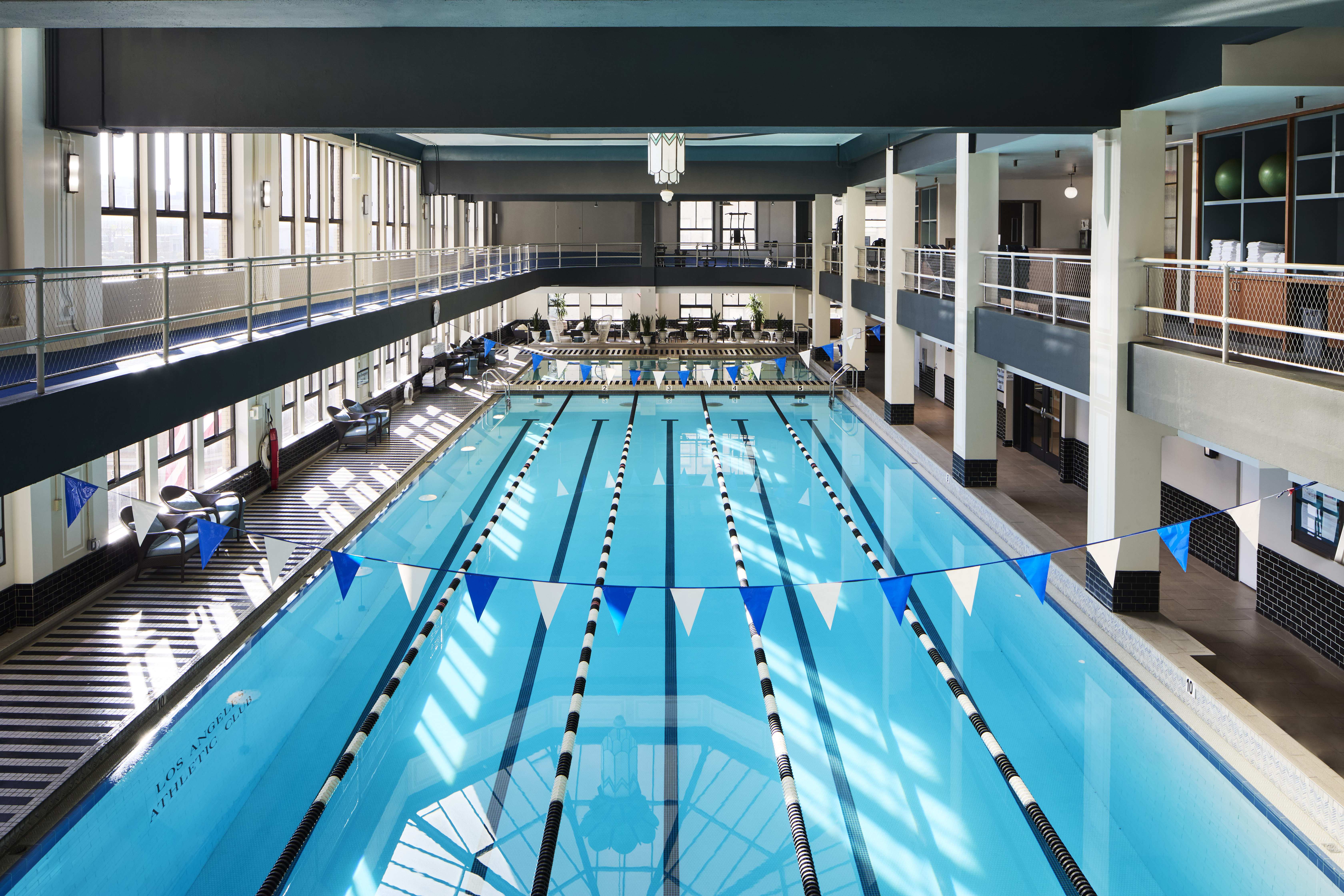 An indoor swimming pool with blue water, black and white lane dividers, and flags across it. There is a balcony area with seating and a lantern hanging from the ceiling.